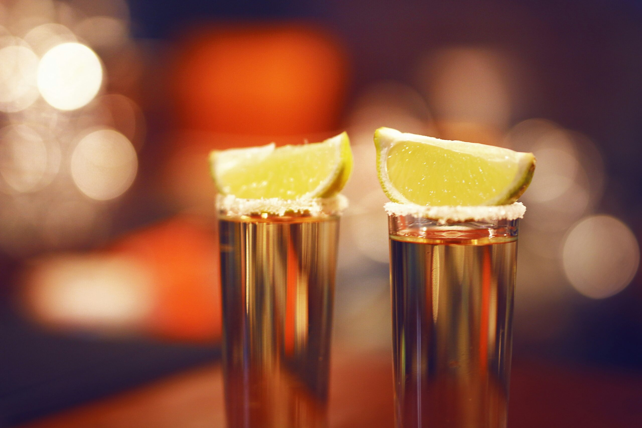 Two tequila shots with lime wedges and salt on the rim, set against a blurred background, creating a festive and vibrant atmosphere