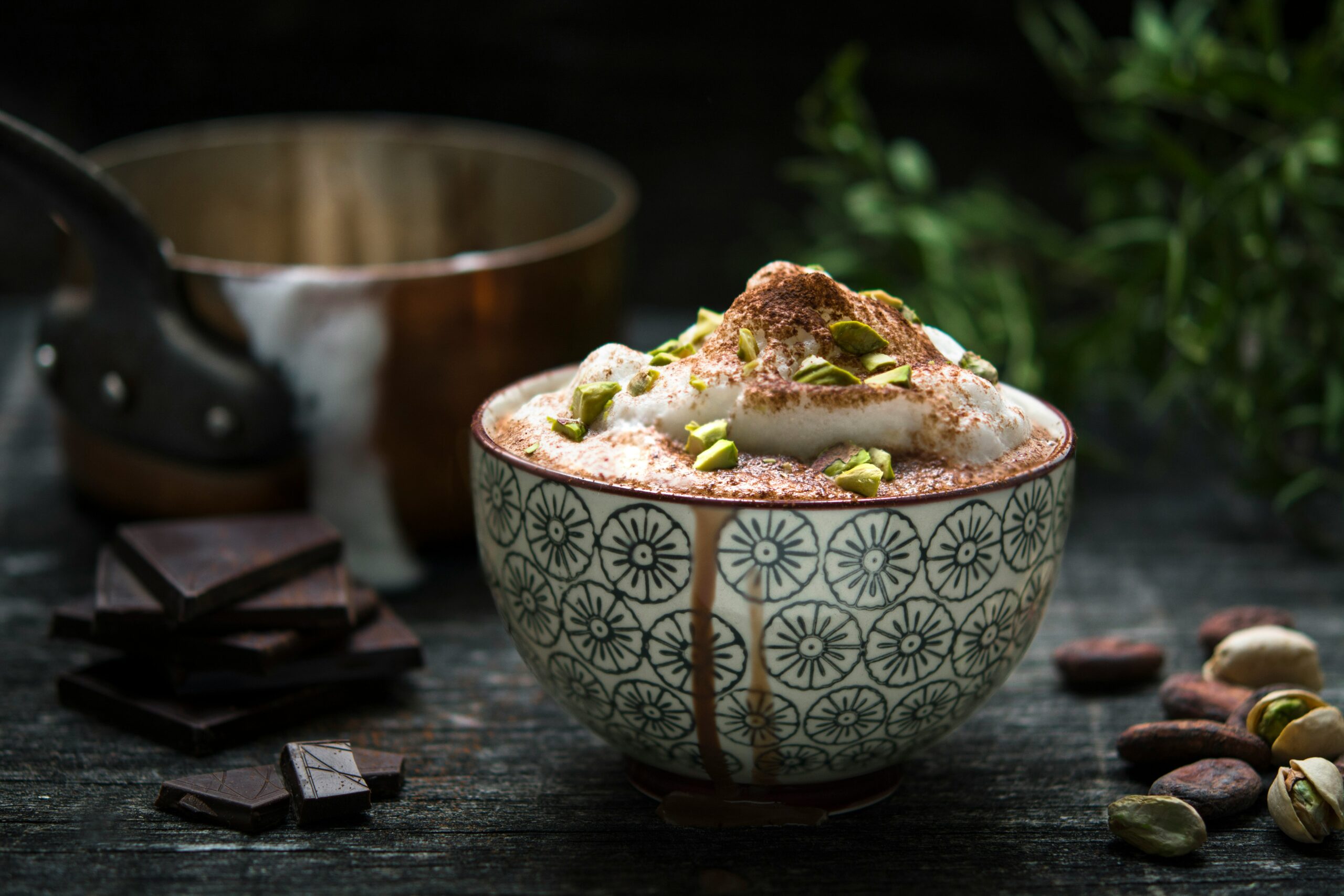 Rich hot chocolate with liquor, topped with whipped cream, cocoa powder, and chopped pistachios in a decorative bowl. The rustic background features dark chocolate pieces, whole pistachios, cocoa beans, and a copper pot, creating a cozy, earthy ambiance