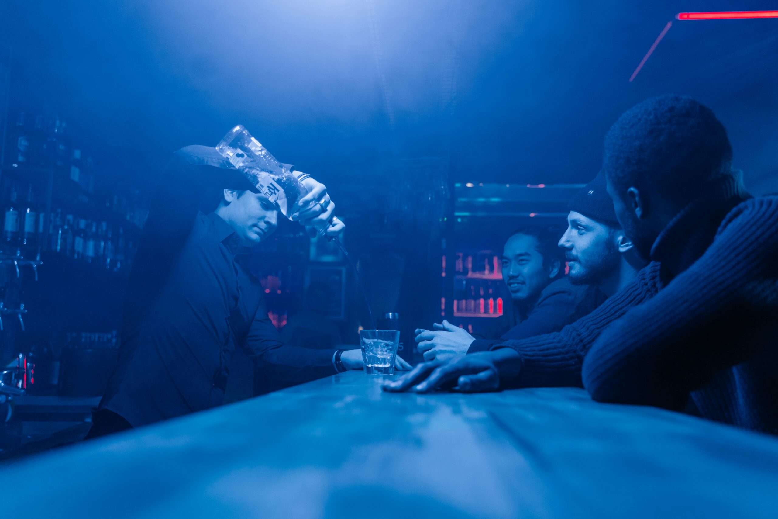 Professional bartender pouring vodka into a glass in a dimly lit bar, with a group of friends chatting in the background under blue ambient lighting